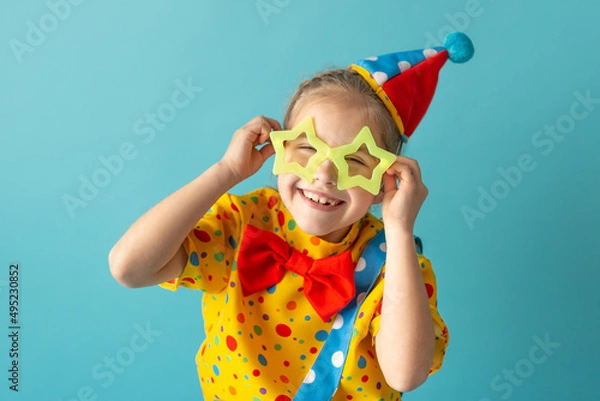 Fototapeta Funny kid clown against blue background. Happy child playing with festive decor. Birthday and 1 April Fool's day concept