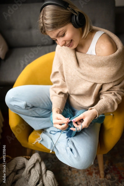 Fototapeta A young smiling girl is sitting in a yellow armchair and knitting a sweater with blue thread