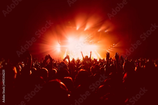 Fototapeta A crowded concert hall with scene stage in red lights, rock show performance, with people silhouette, colourful confetti explosion fired on dance floor air during a concert festival