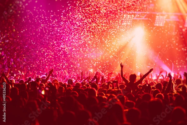 Fototapeta A crowded concert hall with scene stage in red lights, rock show performance, with people silhouette, colourful confetti explosion fired on dance floor air during a concert festival