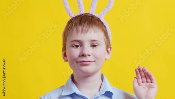 Obraz Close-up of a happy boy with rabbit ears on a yellow background. The boy waves his hand and looks at the camera. Easter