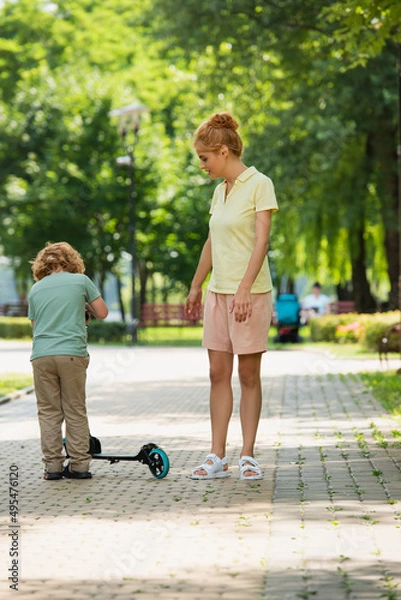 Fototapeta full length view of woman in summer clothes standing near son with kick scooter in park.