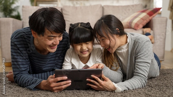 Obraz happy Asian family of three watching funny online video with tablet computer together at home. they lie prone on living room floor and point at screen while laughing