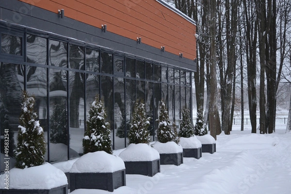 Fototapeta The exterior of a commercial building with large windows in the snowdrifts in winter. The windows reflect the plants planted in front of the building.
