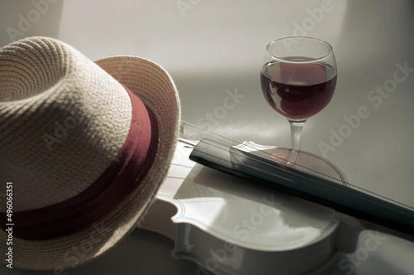 Fototapeta Romantic plot with a white violin and a glass of red wine on a gray background, with a straw hat with a red piping