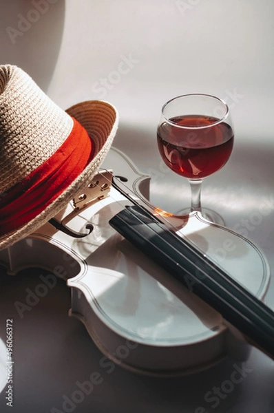 Fototapeta Romantic plot with a white violin and a glass red wine on a gray background, with a straw hat with a red piping
