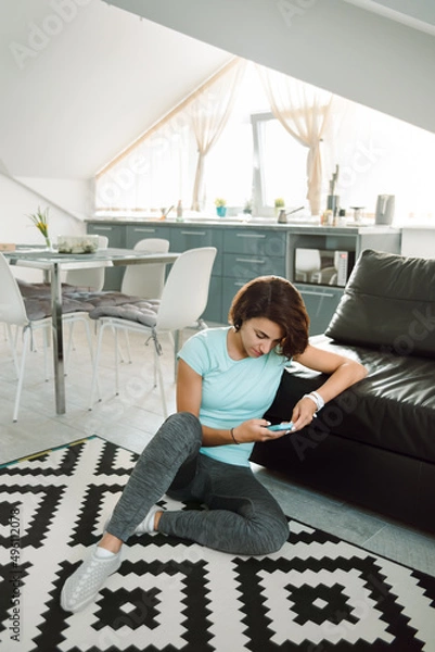 Obraz Young brunette girl sit on the floor in apartment and look at phone .
