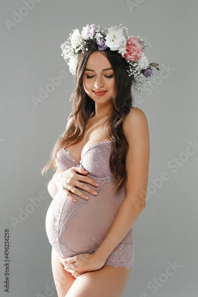 Fototapeta Beautiful smiling pregnant girl in a cute underwear and a wreath of flowers on her head posing in a studio