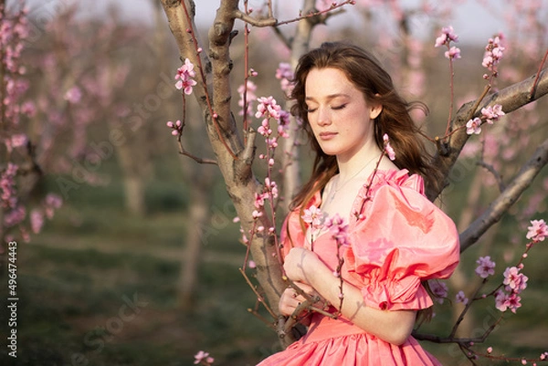 Fototapeta Portrait of a young girl in peach orchards. Blooming trees in spring and a beautiful girl in a pink dress stands with her eyes closed.