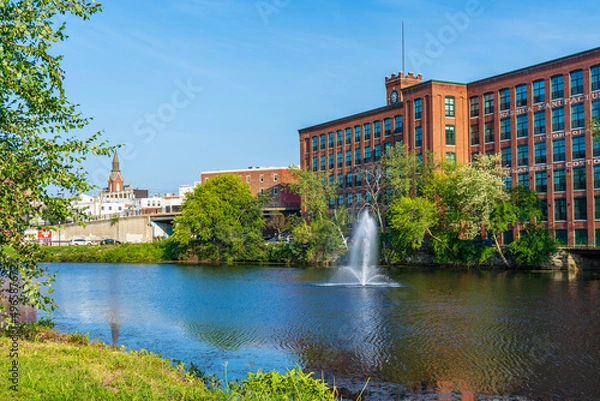 Fototapeta Fountain on the Nashua River against the background of a historic cotton factory building with a clock tower in the old industrial park of Nashua. New Hampshire, USA