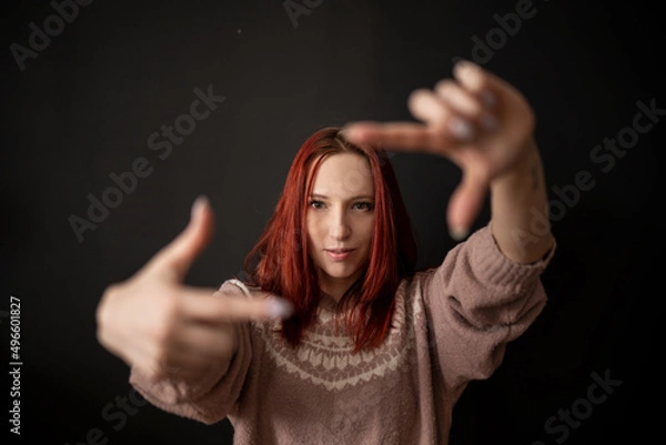 Fototapeta a beautiful and natural smiling red-haired skinny girl in a knitted sweater makes an imaginary lens or camera frame gesture with her fingers and looks through it at the viewer
