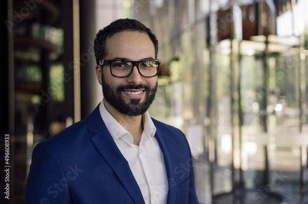 Fototapeta Confident smiling Arabian business man wearing stylish eyeglasses looking at camera standing in modern office. Portrait of handsome Moroccan manager at workplace.  Successful business concept