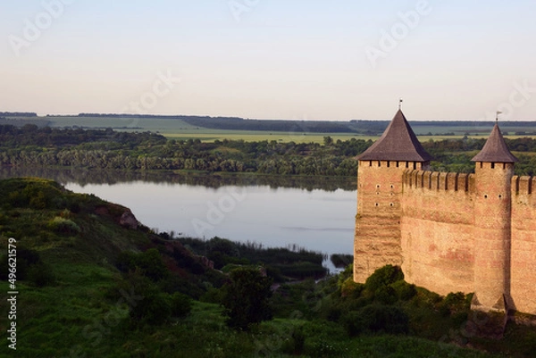 Fototapeta On the right side of the tower of a medieval stone fortress with battlements. On the left is a river with banks in the grass