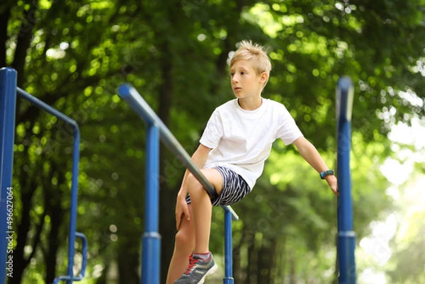 Fototapeta a boy on a sports ground in a park climbed onto a sports equipment and looks while sitting on it