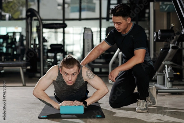 Fototapeta Fit young man doing push-up exercise under control of his fitness trainer in gym