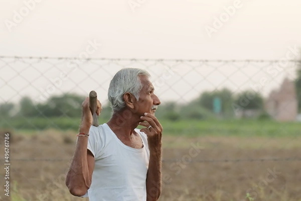 Fototapeta An Indian senior aged man farmer thinking about something while holding a shovel in his hand at sunset