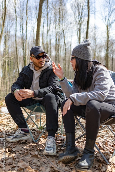 Fototapeta Young man and woman sitting on chairs in forest and having a conversation. Friendship, joy, social life and smiling concept. They are drinking tea. Blur trees background. Vertical photo.