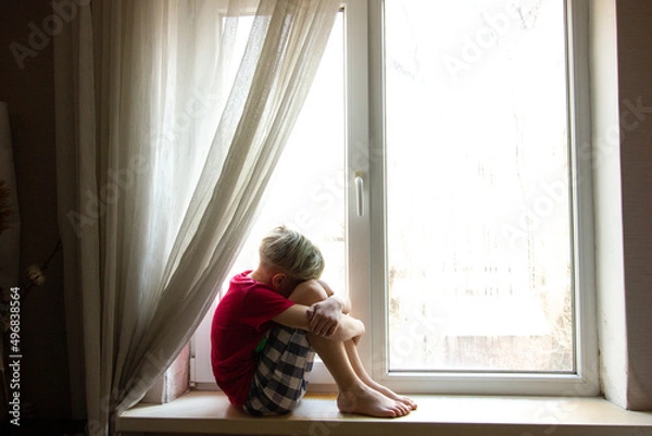 Fototapeta Sad teenager boy sitting on the sill of the large window in a dark room and looks forward.