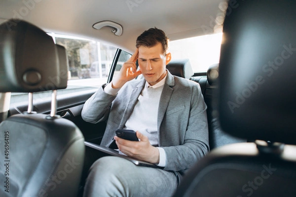 Fototapeta Young businessman tired from work holding hand to his head while sitting in the back seat of the car.