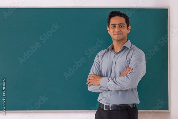 Fototapeta School teacher standing in front of blackboard with arms folded in classroom