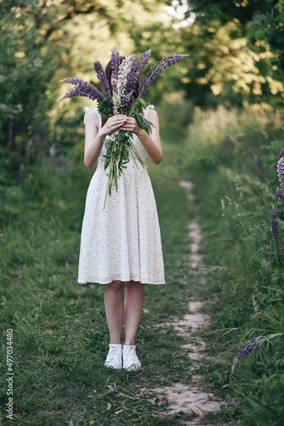 Obraz Young girl covers her face with lupine flowers in warm spring in a green park