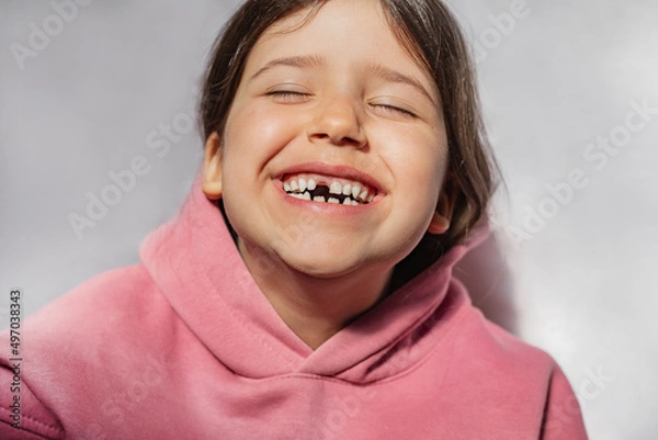 Fototapeta portrait of a beautiful emotional caucasian girl in a pink sweatshirt who lost her first tooth. Close-up of a child with a smile and joy