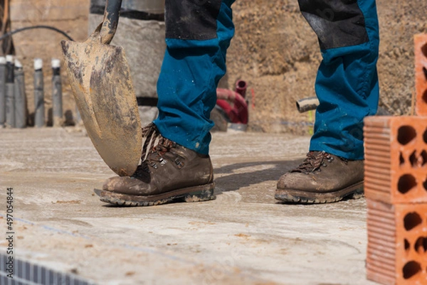 Fototapeta Worker's legs in construction site with a shovel on safety boots and bricks