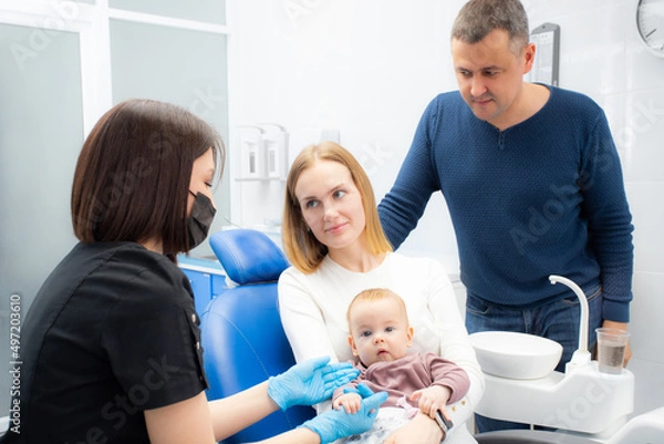 Obraz Young parents and their daughter visit a dentist to examine milk teeth. Baby at the first appointment with the dentist. Inspection of the formation of jaws, eruption of milk teeth.
