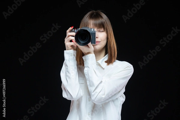 Fototapeta Portrait of a young woman wearing a white shirt holding a camera in her hand. The concept of a successful photographer. Isolated black background.