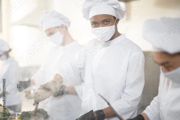 Fototapeta Multiracial team of chef cooks in white uniform cooking together in the kitchen. Asian chef with latin cook frying meat, european guys cooking on background. Cooks wearing face masks and protective