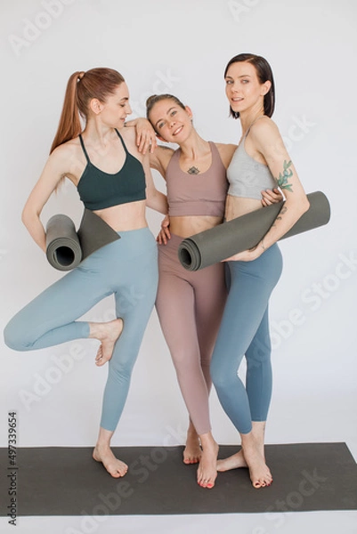 Fototapeta three beautiful girls smiling, hold yoga mats in their hands and 
look at each other in a white studio. Girl doing tree pose
models are dressed in sports leggings and top.     