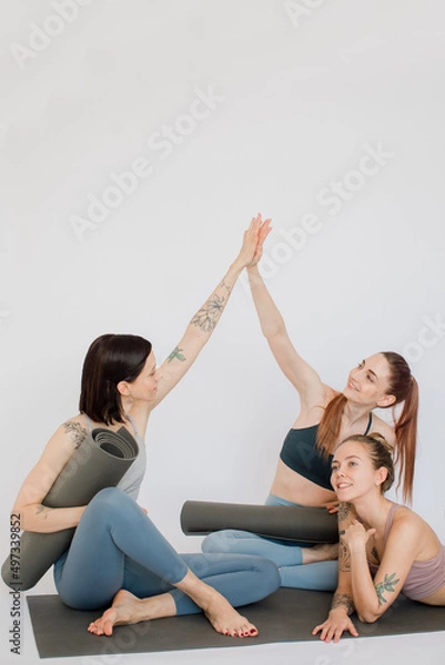 Fototapeta three beautiful girls smile, relax sitting on a yoga mat, make a handshake, joyfully give five in a white studio.
models are dressed in sports leggings and tops