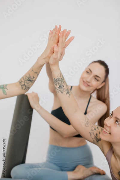 Fototapeta three beautiful girls smile, relax sitting on a yoga mat, make a handshake, joyfully give five in a white studio.
