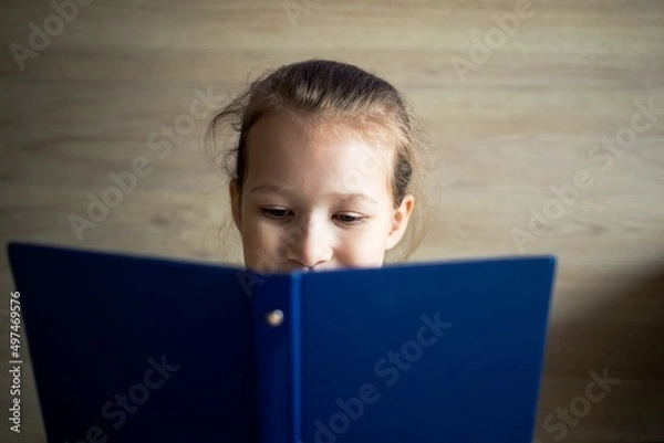 Fototapeta little girl child reads a blue book and looks out from behind it, eyes and a smile are visible, school tomorrow