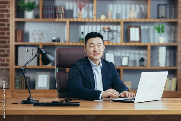 Fototapeta Portrait of a successful Asian businessman, man working in the office sitting at the table, looking at the camera and smiling, happy banker with laptop