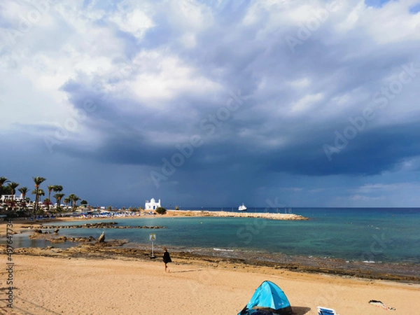 Fototapeta The sunny sandy coast of the Mediterranean Sea, the beach in the bay, the temple of St. Nicholas the Wonderworker, a dramatic sky and rain over the sea, a ship in the sea.