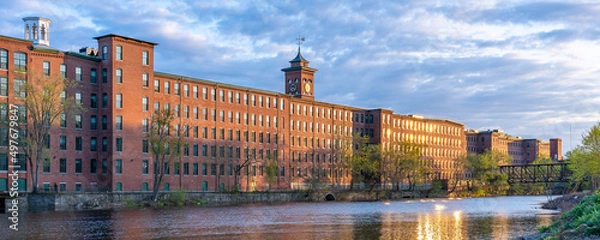 Fototapeta Historic cotton mill building with clock tower in an old industrial park on the Nashua River illuminated by the sun during sunset in May. Panoramic photography. Nashua, New Hampshire, USA