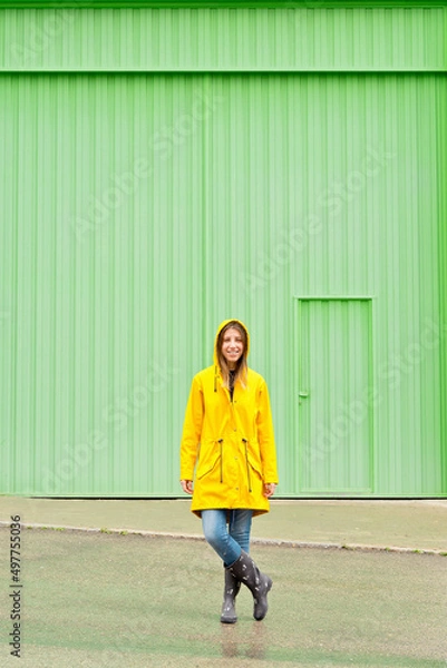 Fototapeta Vertical front view of woman wearing a yellow raincoat standing on the street. Full length body of woman under the rain with wellingtons and green facade. People isolated in background concept.