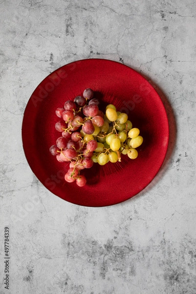 Fototapeta Still life dark and green grapes in on a red dish in a stone background