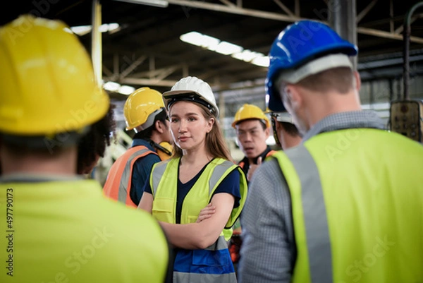 Obraz A confident, beautiful Caucasian woman worker stands among the male workers wearing safety uniform are meeting morning talk before the start of work in heavy industrial manufacturer factory.