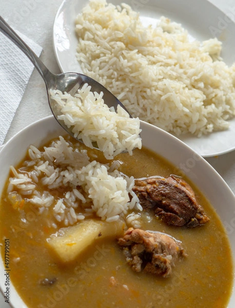 Fototapeta A white bowl of Dominican sancocho with yucca, chicken, pork, squash and more, served with white rice and a metal spoon.