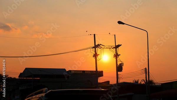 Obraz Silhouettes buildings, high voltage poles, streetlight poles, power lines and birds flying in the golden sky during sunrise or sunset.