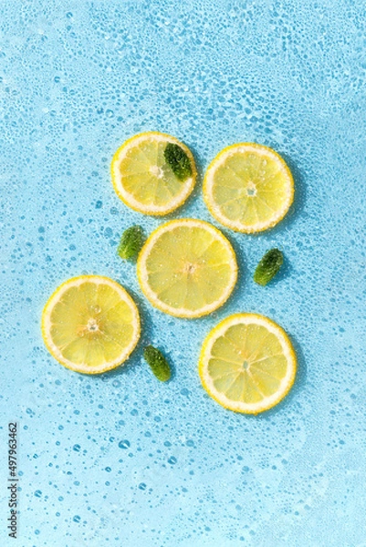 Fototapeta Round lemon slices in water with bubbles and green leaves. Cut citrus in carbonated blue soda. Concept of summer refreshing.