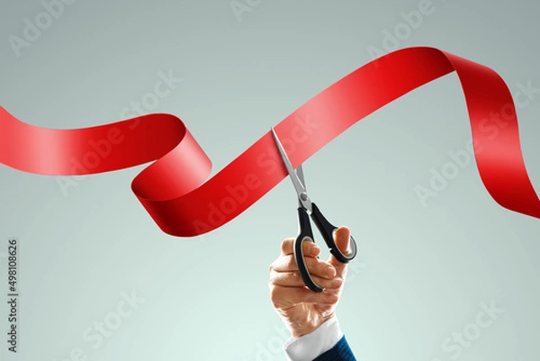 Fototapeta Grand opening with red ribbon and scissors. A businessman's hand holds scissors cuts a red ribbon on a light background. Close-up, copy space.