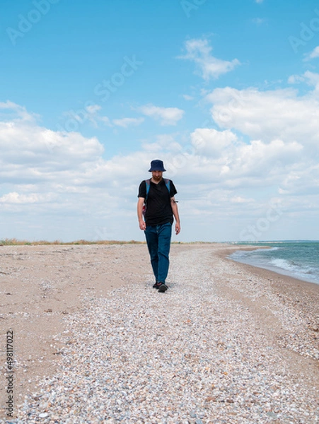 Fototapeta Smiling bearded millennial man in bucket hat black t-shirt walking on empty beach. Authentic male tourist lifestyle photo. Hipster guy outdoor. Solo travel adventure concept Active walking backpacking