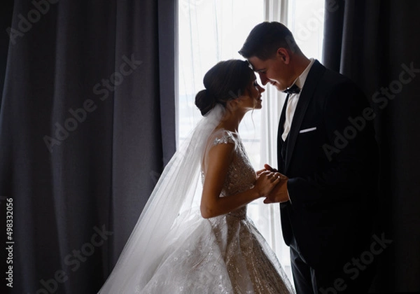 Obraz Side view of adorable couple in festival outfit, standing face to face in room, closing eyes, holding hands and feeling happy during wedding day