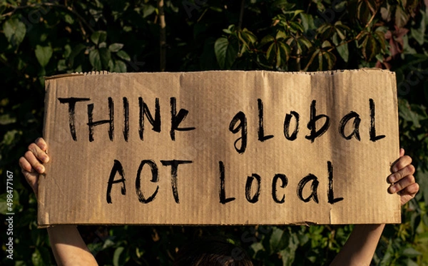 Fototapeta A poster with the words THINK GLOBAL, ACT LOCAL in the hands of a young girl on a background of green foliage.