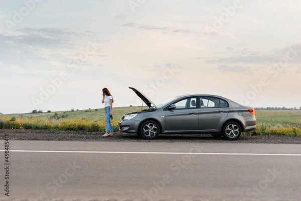 Fototapeta A young girl stands near a broken car in the middle of the highway during sunset and tries to call for help on the phone and start the car. Waiting for help. Car service. Car breakdown on the road.