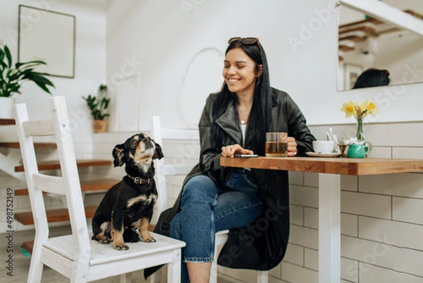 Obraz An optimistic woman in a gray suit smiles and plays with a dog in a cafe. Pretty woman in stylish coat posing