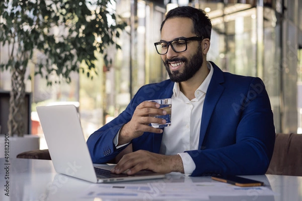 Fototapeta Handsome smiling Moroccan businessman using laptop computer working online sitting in modern office. Portrait of happy successful manager drinking water planning project sitting at workplace
